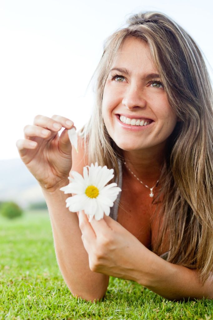 A woman with a look of adoration and a smile, holds up a daisy while plucking at its leaves